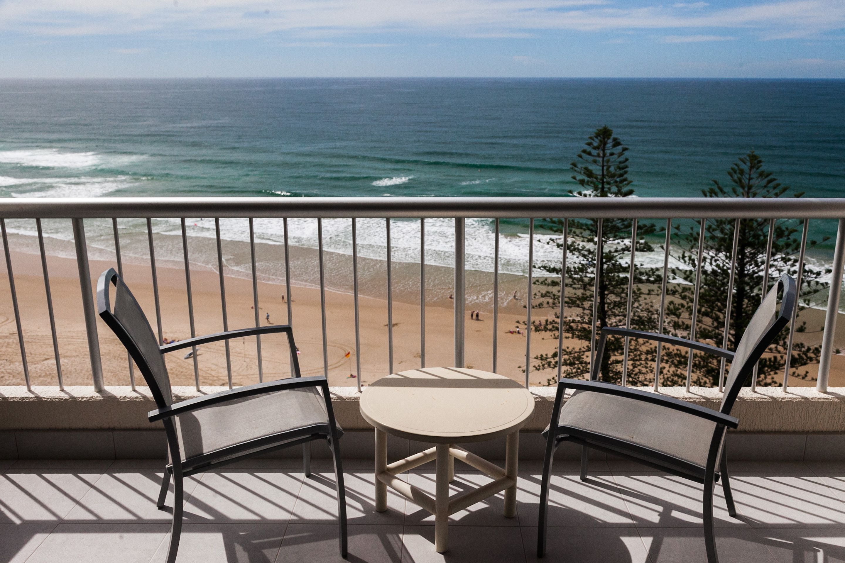 A balcony with two chairs and a small table, overlooking a beach and the ocean.