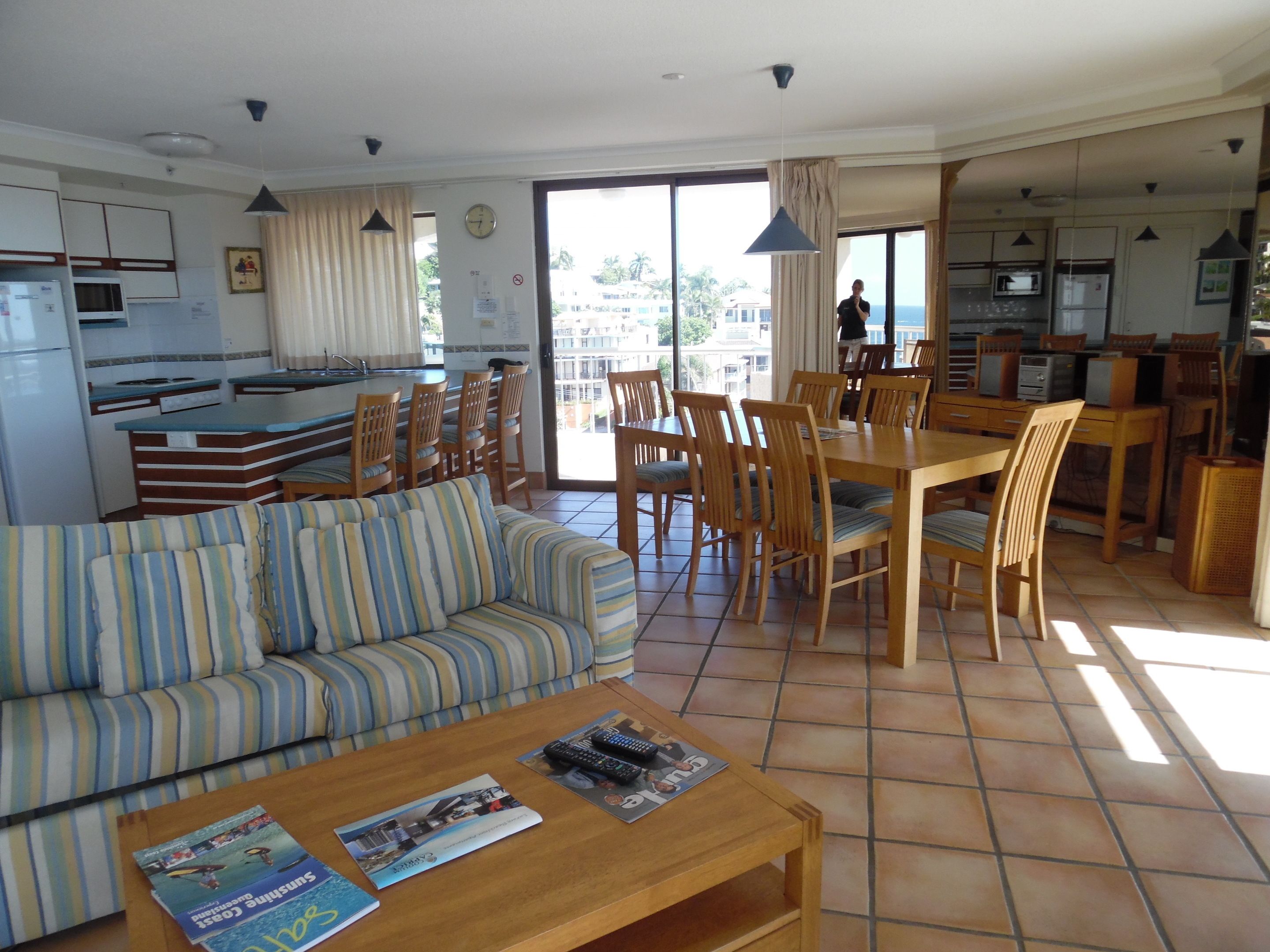 A spacious living area with a sofa, dining table, and chairs, a kitchen with a refrigerator and microwave, and large windows offering a view of the surroundings.
