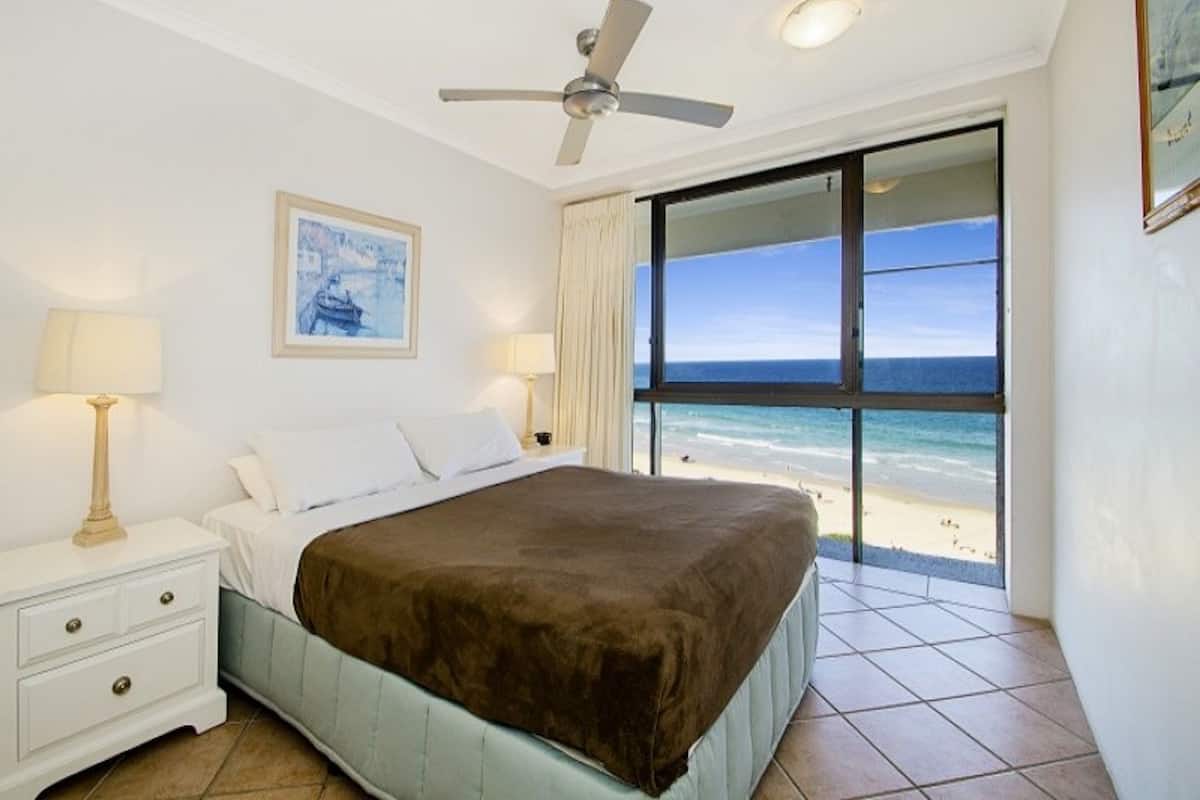 A hotel room with a bed, a nightstand, a ceiling fan, a window with a beach view, and a framed picture on the wall.