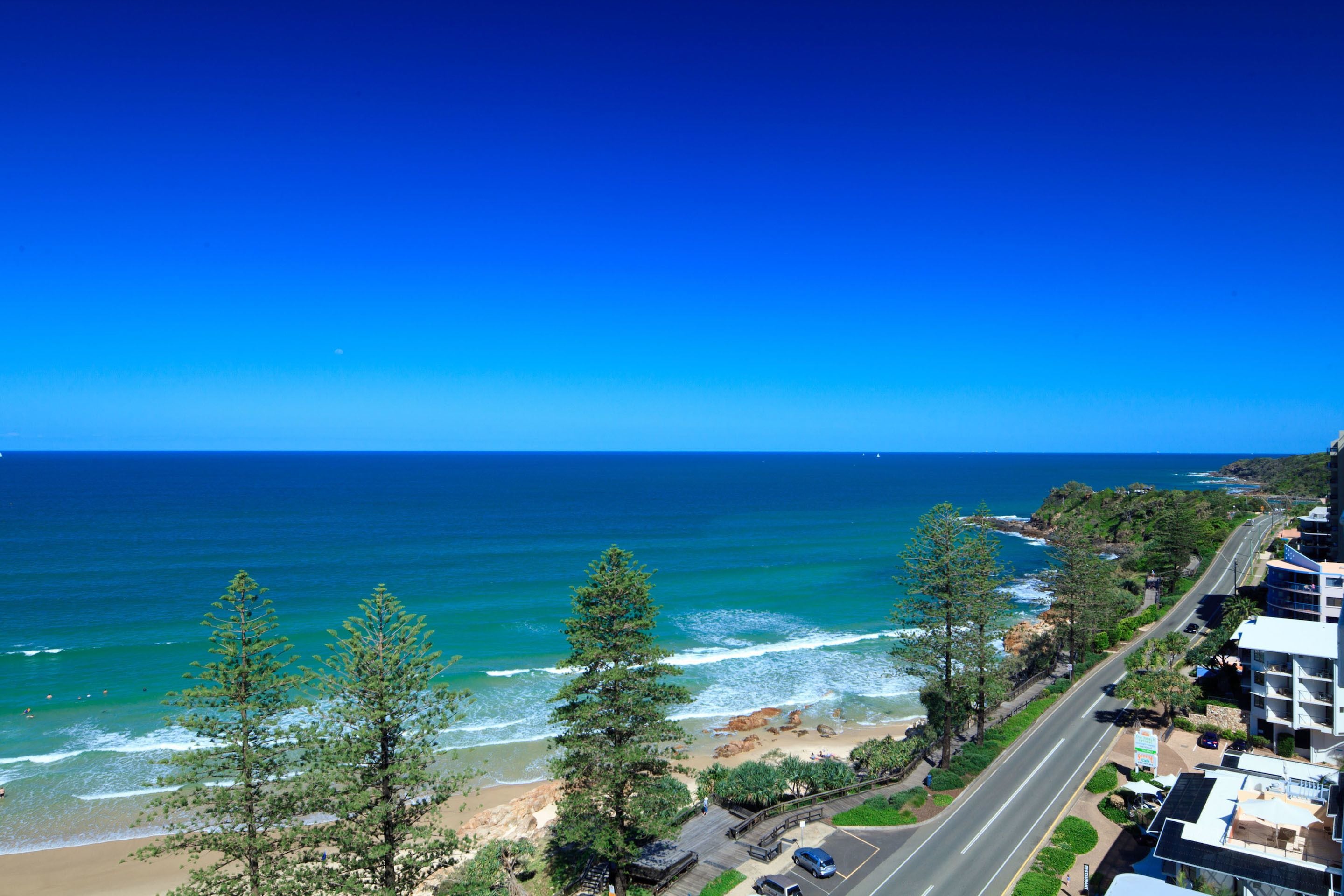 A coastal view with a road leading to a beach, trees, and a building with balconies overlooking the ocean.
