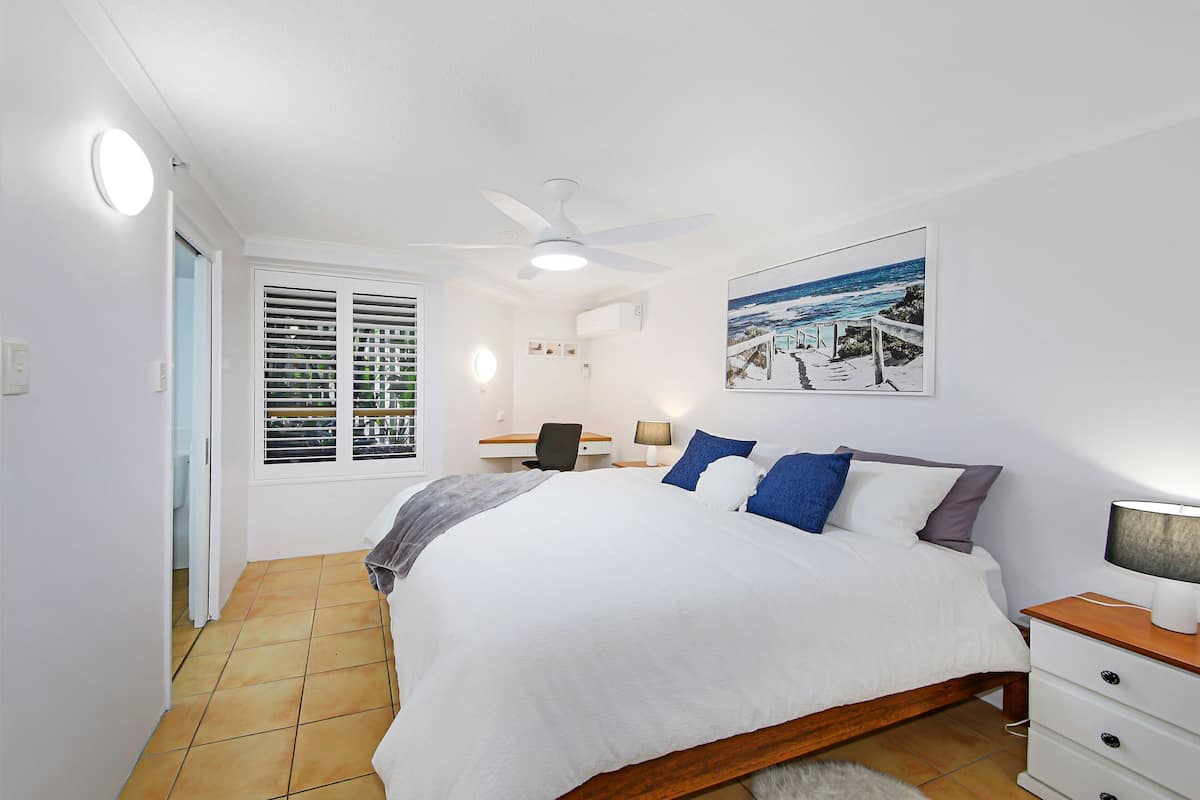 A bedroom with a bed, a desk, a chair, a ceiling fan, a window with blinds, and a painting on the wall.