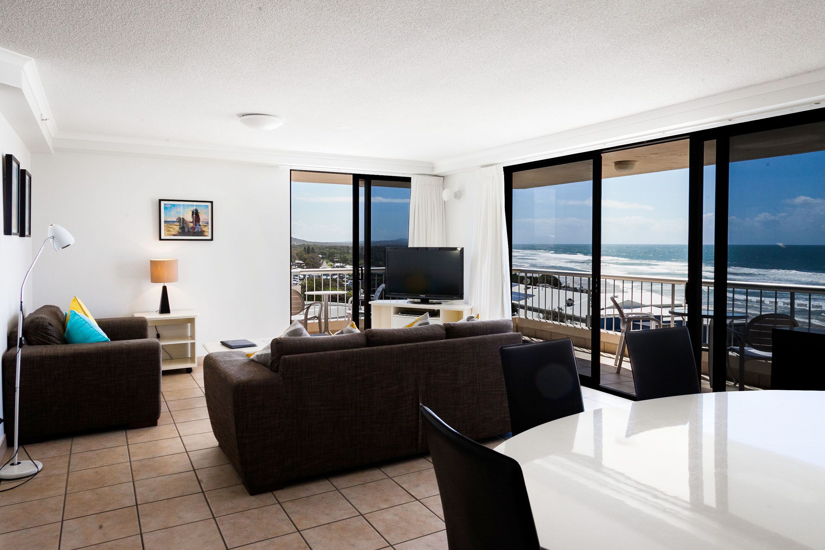 A modern living room with a large TV, comfortable sofas, a dining table, and a balcony with a view of the ocean.
