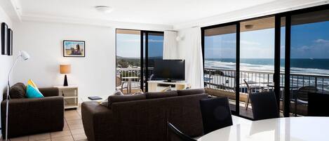 A modern living room with a large TV, comfortable sofas, a dining table, and a balcony with a view of the ocean.