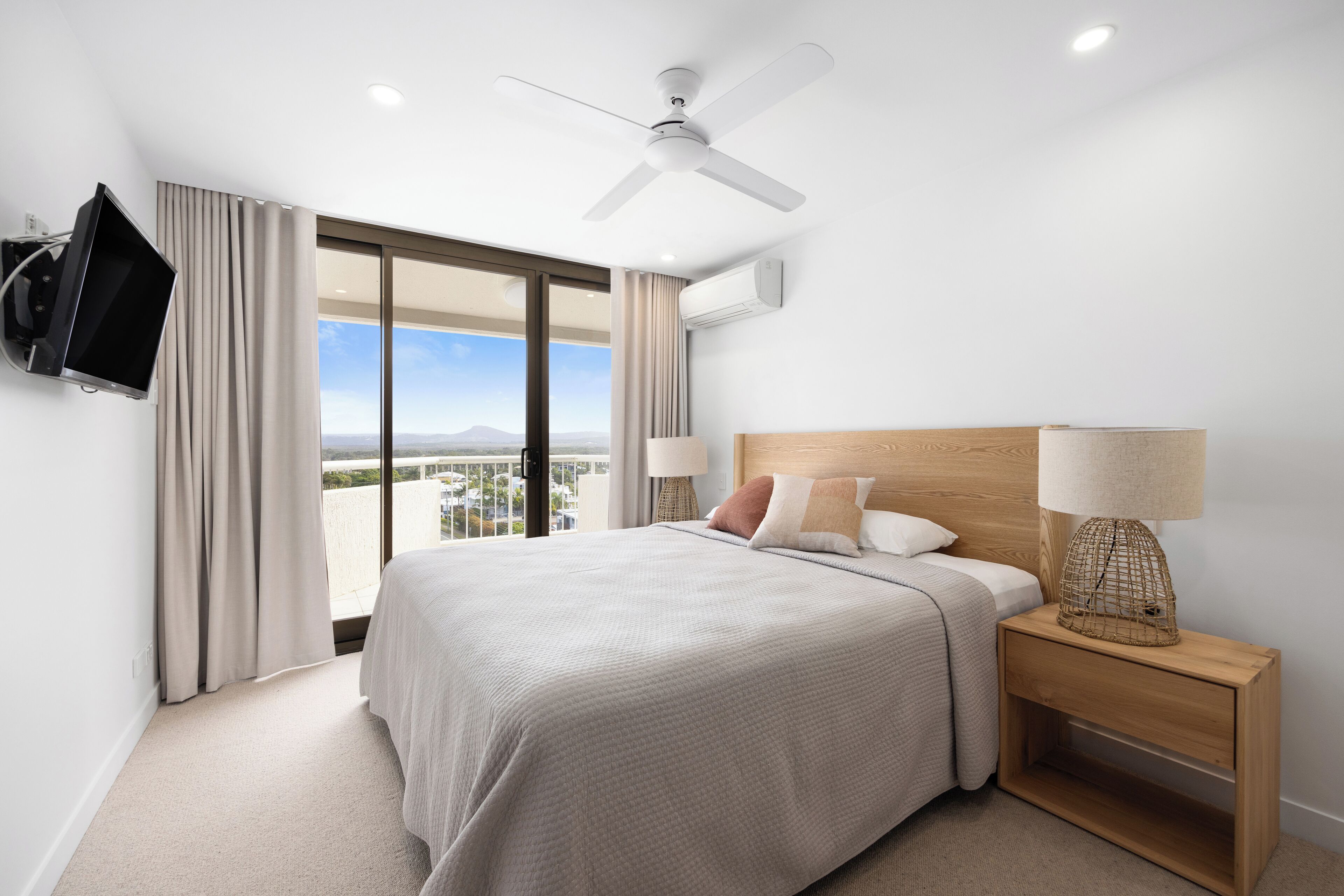 Une chambre d’hôtel moderne dotée d’un grand lit, d’un ventilateur de plafond, d’un téléviseur fixé au mur, d’une table de chevet avec une lampe et offrant une vue sur la ville grâce à de grandes fenêtres.