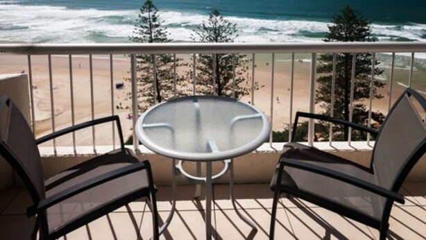 A balcony with a glass table and two chairs, overlooking a beach with waves and trees. - Coolum Caprice (Coolum Beach)