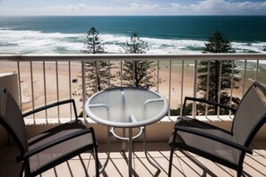A balcony with a glass table and two chairs, overlooking a beach with waves and trees. - Coolum Caprice (Coolum Beach)