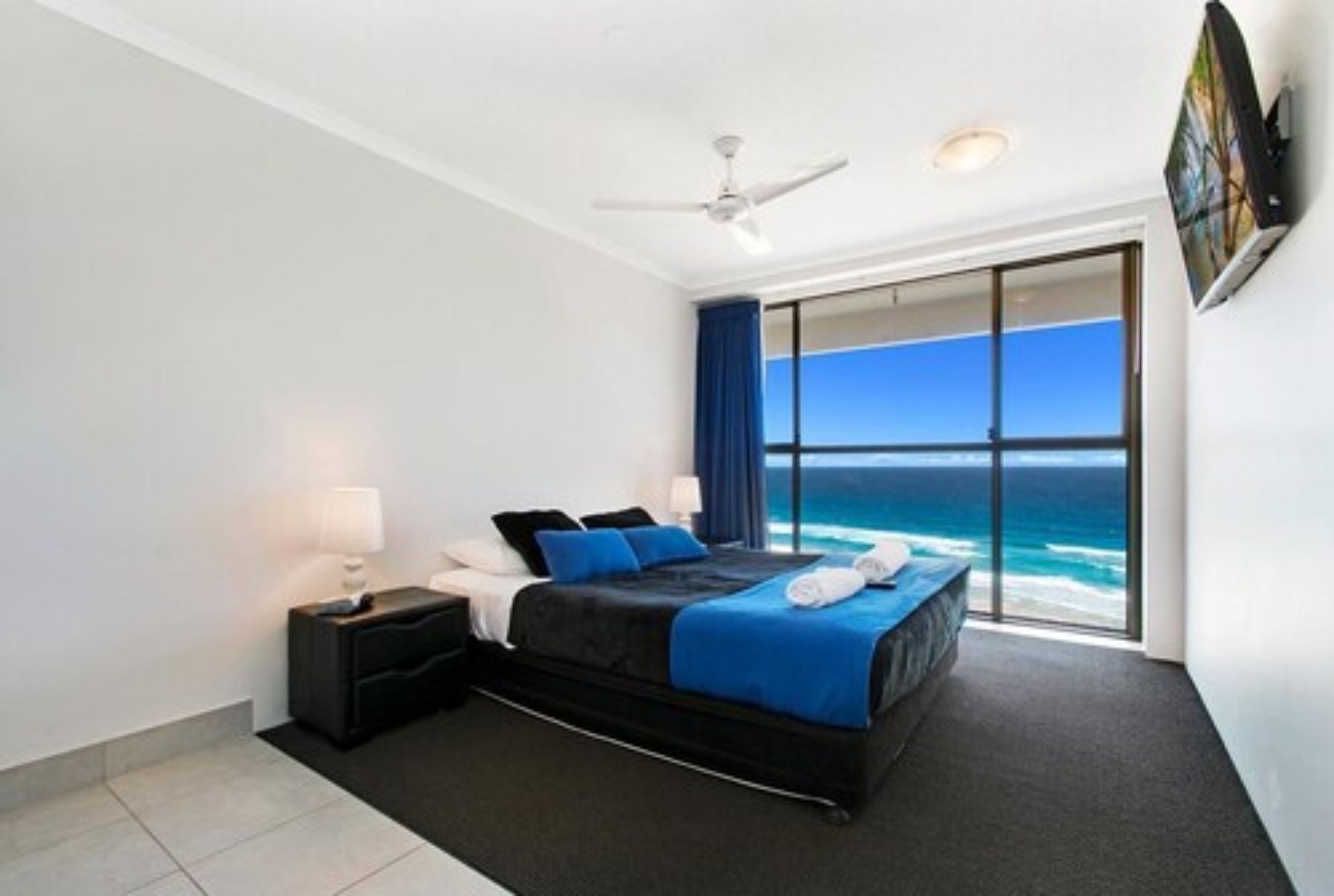 A bedroom with a bed, a nightstand, a ceiling fan, and a large window with a view of the ocean.