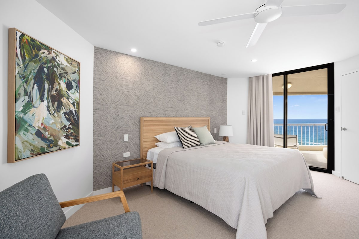 A modern hotel room with a large bed, a nightstand, a chair, a ceiling fan, and a view of the sea through a sliding door.