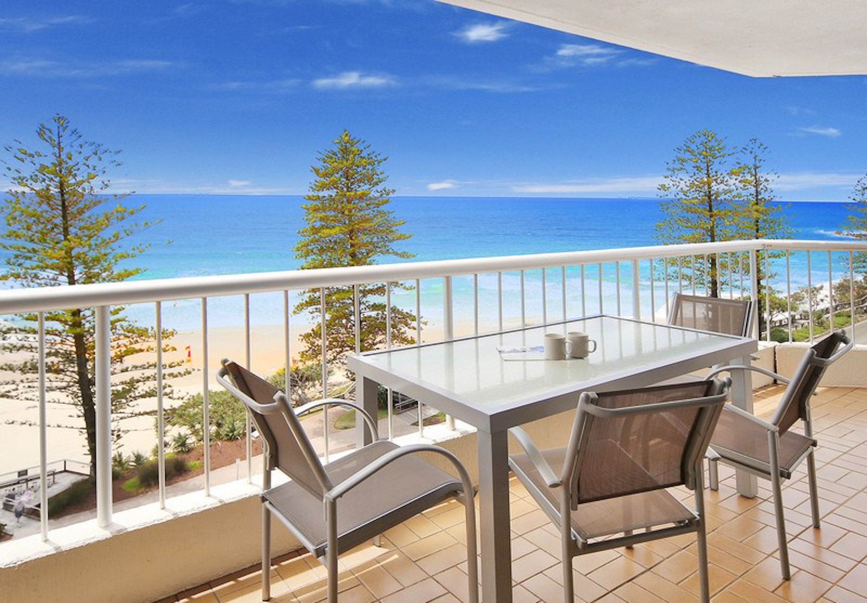 A balcony with a glass table and chairs, overlooking a beach and the sea.