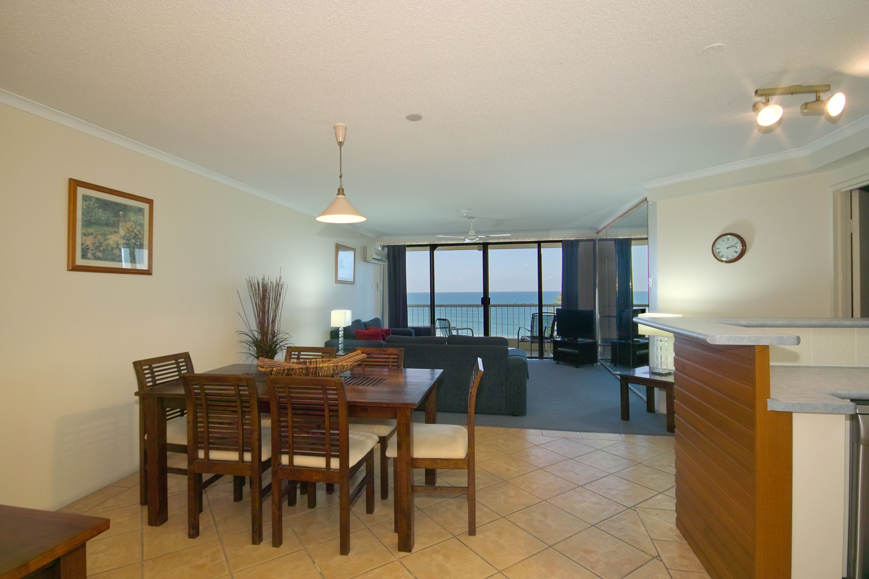 A dining area with a wooden table and chairs, a kitchen counter, and a view of the ocean through large windows.