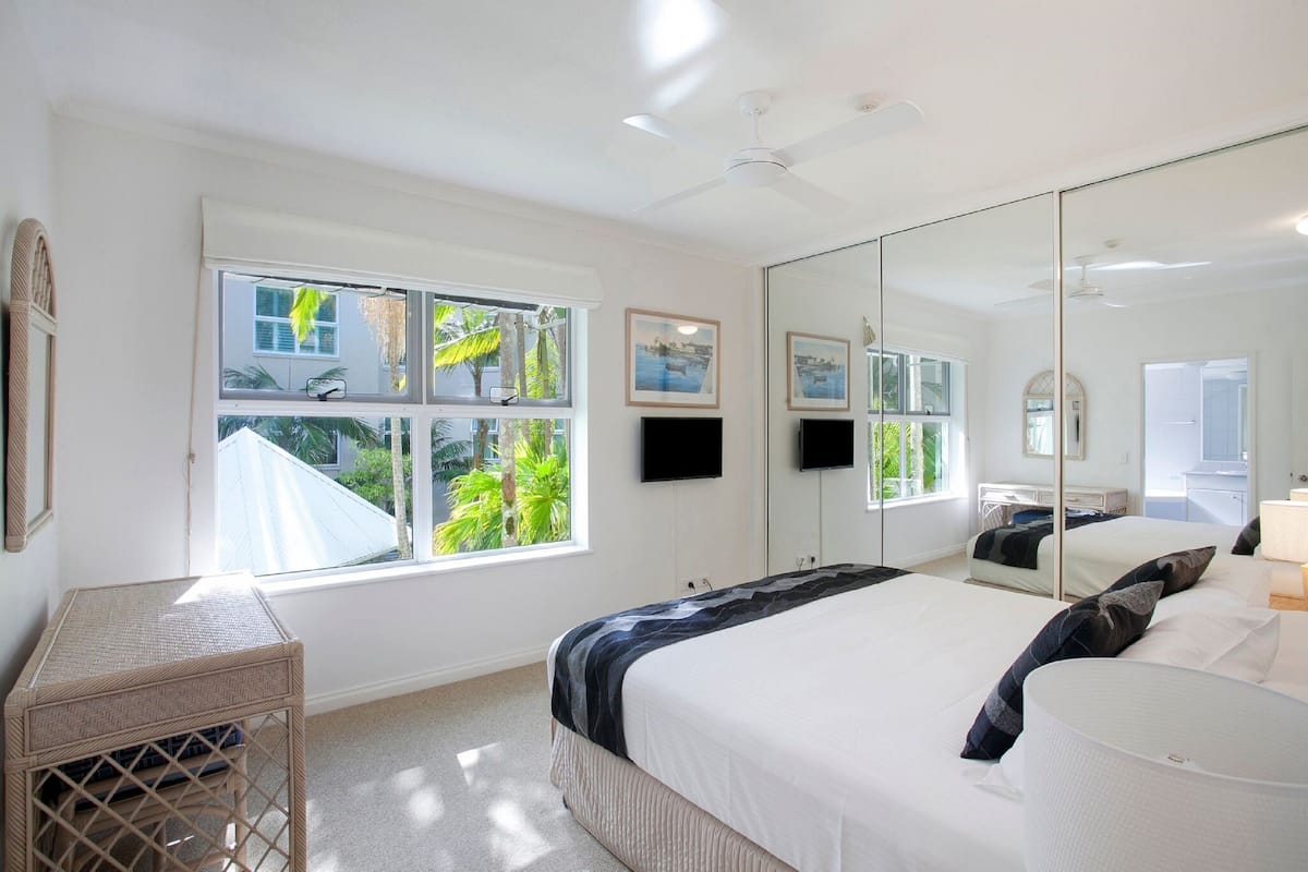 A bedroom with a large bed, a desk, a mirror, and a window with a view of greenery.