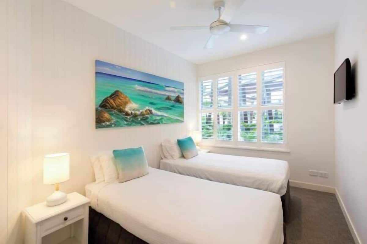 A hotel room with two beds, a painting of a coastal scene, a ceiling fan, and a TV on the wall.