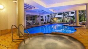 Indendørs swimmingpool med jacuzzi, omgivet af liggestole og palmer.