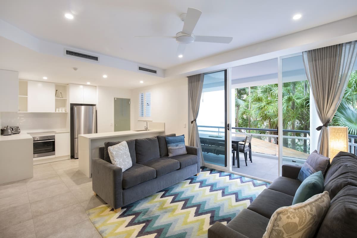 A modern living room with a sofa, a coffee table, and a dining area with a table and chairs. There is a kitchen area with a refrigerator and a stove. The room has large windows and a view of greenery outside.