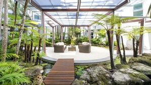 Et overdækket udendørs opholdsområde med træterrasse, omgivet af tropiske planter og sten.