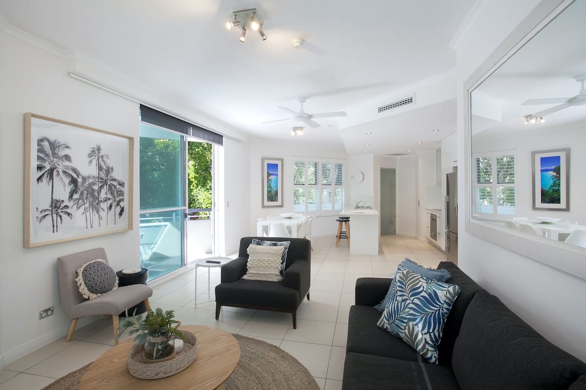 A spacious living area with a modern design, featuring a large window, a black sofa, a grey armchair, and a dining area in the background.