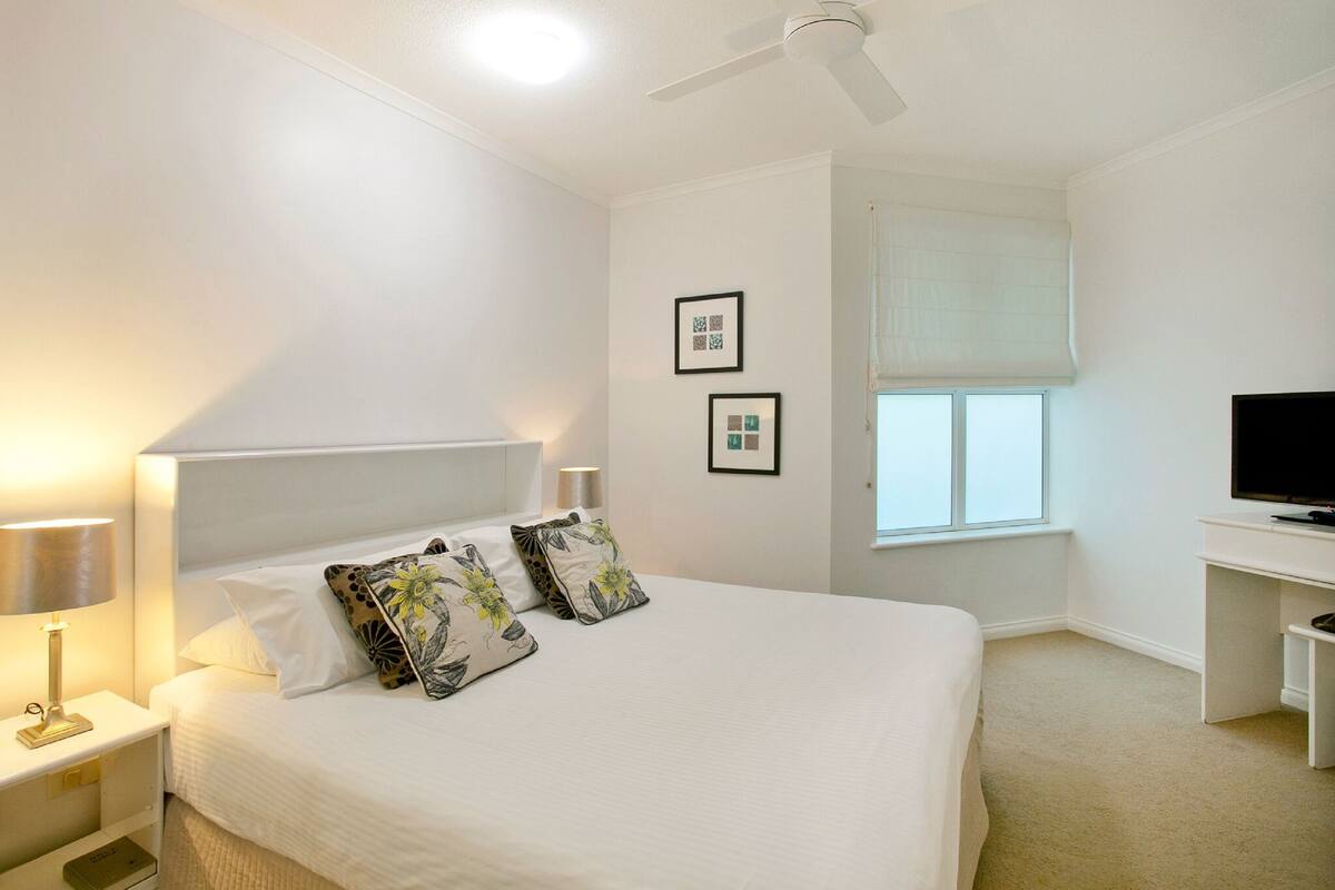 A bedroom with a bed, bedside lamps, a ceiling fan, a TV, and a window with a view.