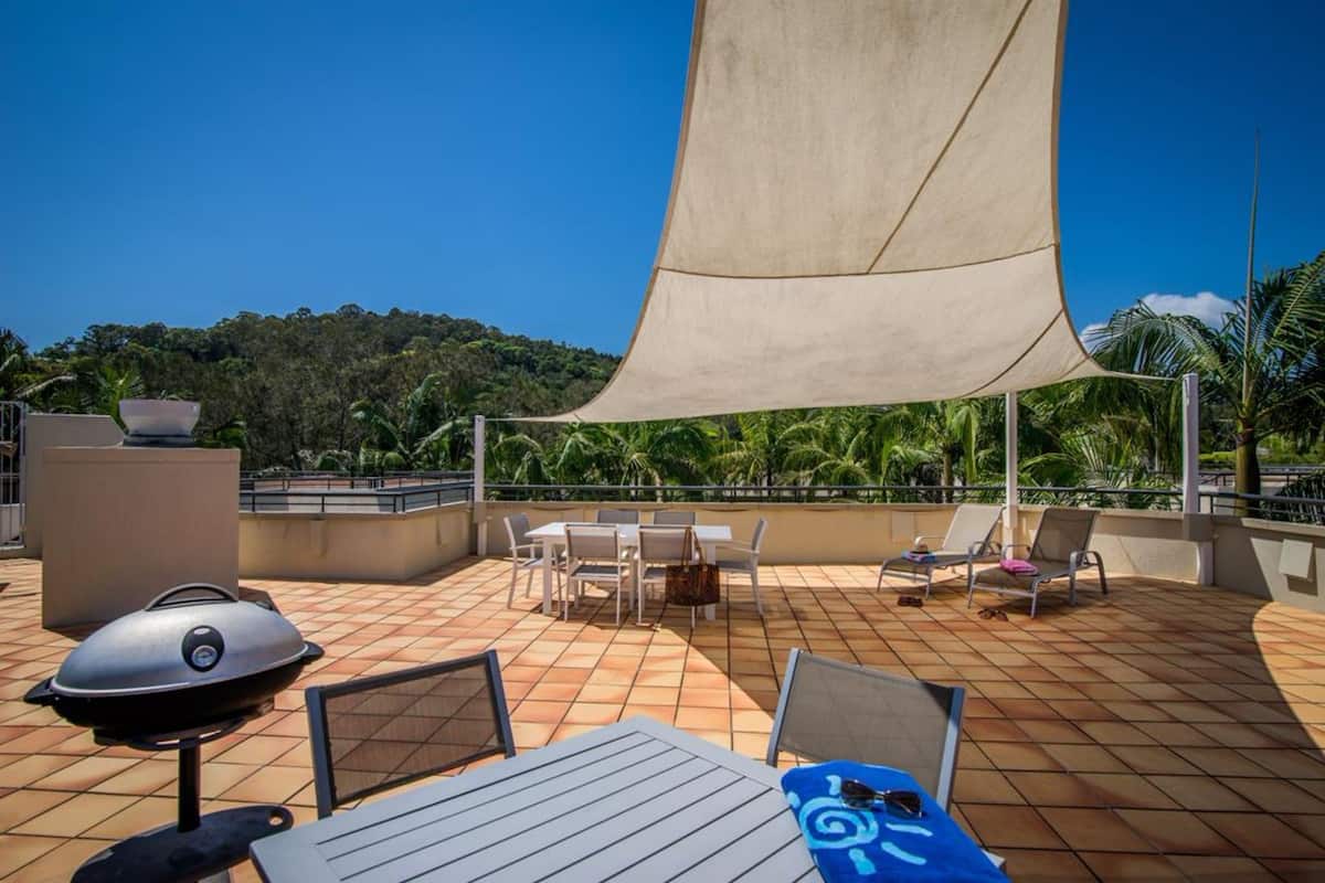 A rooftop terrace with a grill, tables, chairs, and a large shade sail.