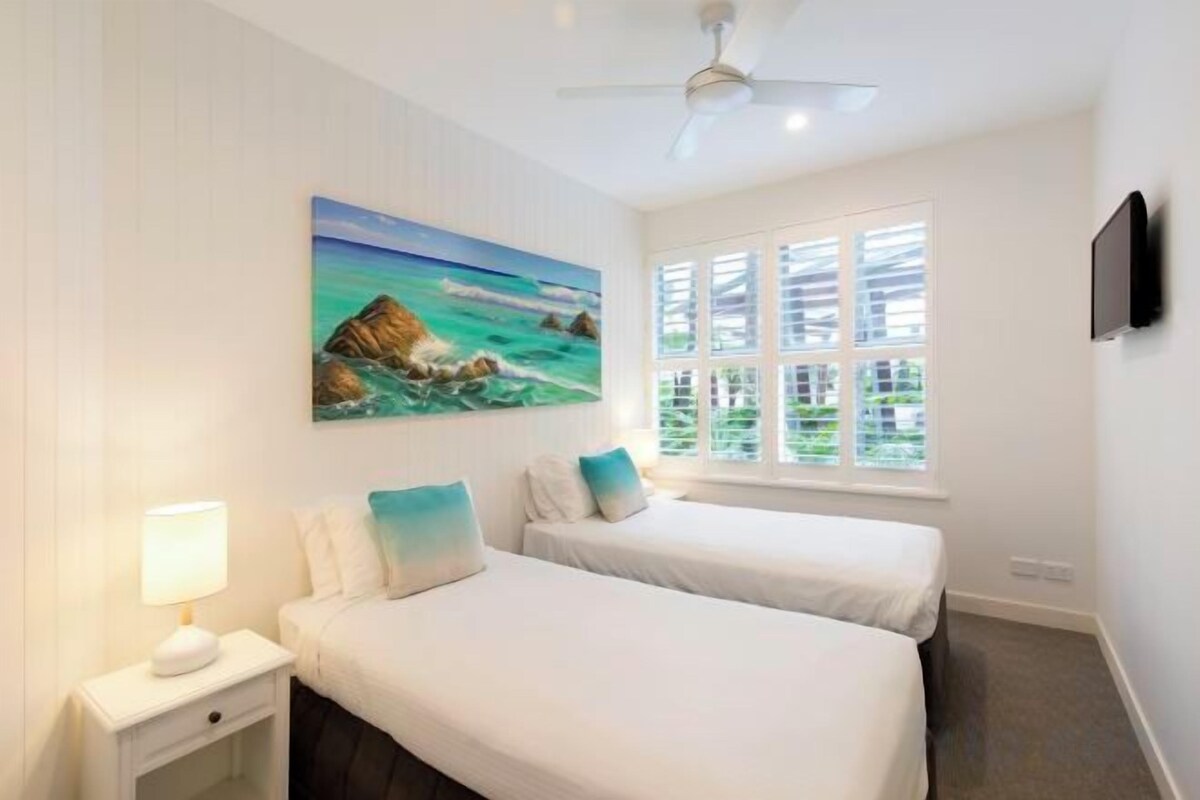 A hotel room with two beds, a painting of a beach scene, a ceiling fan, and a window with blinds.
