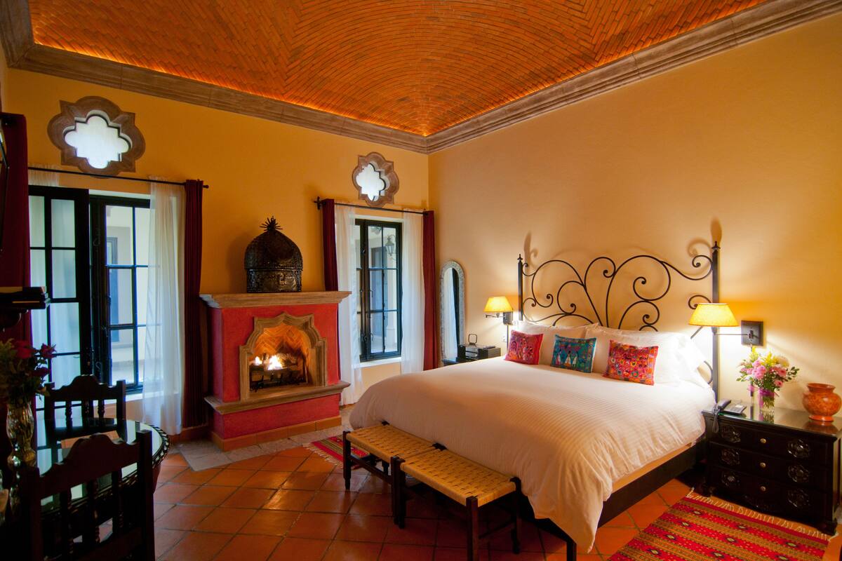 A bedroom with a large bed, a fireplace, a dining table with chairs, and a bench.