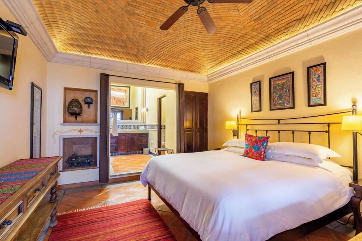 A hotel room with a large bed, a ceiling fan, a fireplace, and artwork on the walls.