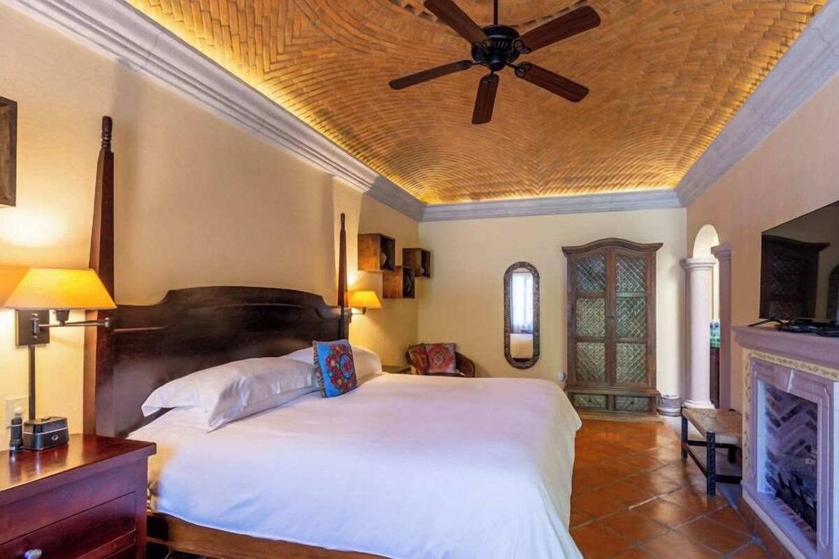 A hotel room with a large bed, a ceiling fan, a fireplace, and a mirror.