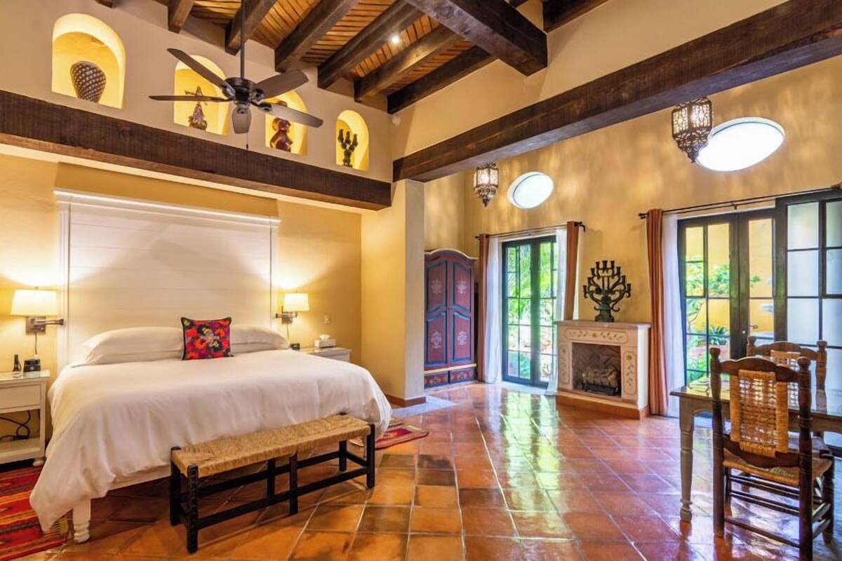 A spacious bedroom with a large bed, a bench, a fireplace, and a dining area with chairs.
