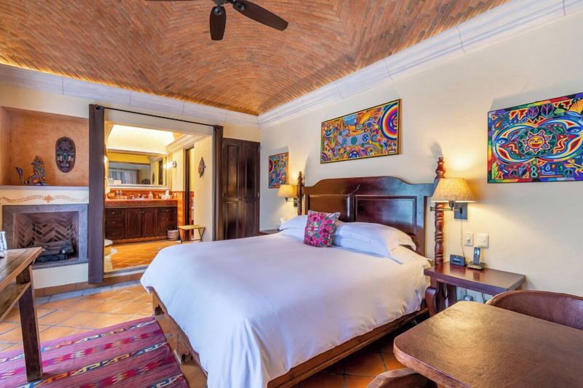 A bedroom with a large bed, wooden headboard, and a ceiling fan. There are colorful paintings on the walls and a fireplace in the corner.