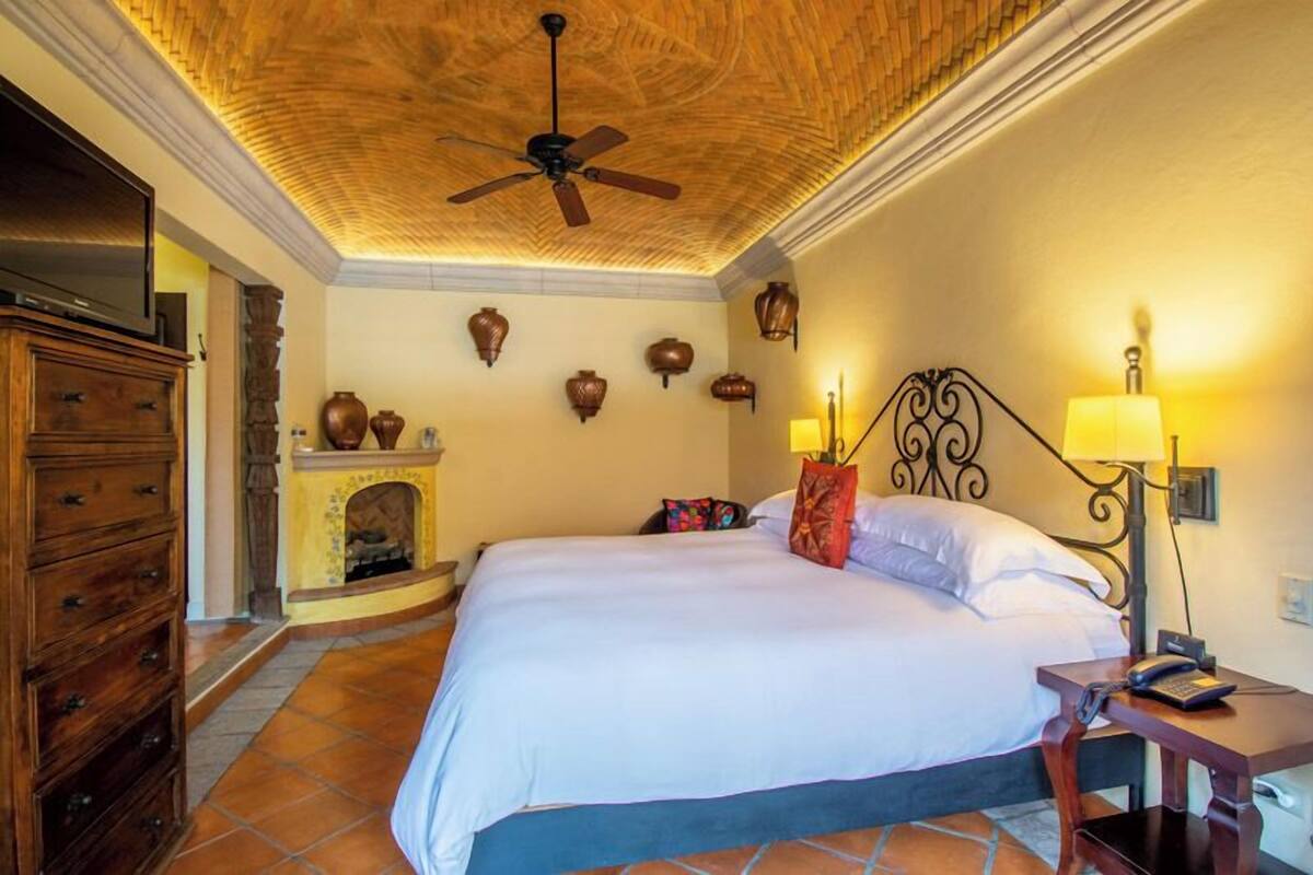 A hotel room with a bed, a wooden dresser, a fireplace, and a ceiling fan.