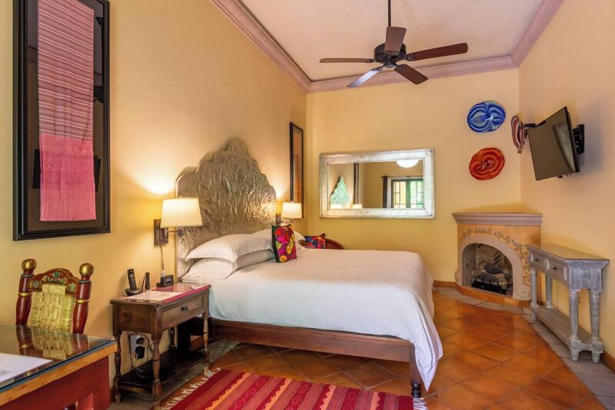 A hotel room with a bed, bedside lamps, a wooden headboard, a desk with a chair, a mirror, a fireplace, and a ceiling fan.