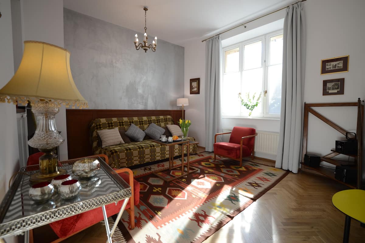 Une pièce bien éclairée, avec un canapé, une table à manger surmontée d’une lampe, une chaise rouge, un tapis à motifs et une fenêtre avec des rideaux.