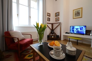 A living room with a red armchair, a coffee table with a vase of yellow flowers, a plate with oranges, and a television displaying a sports game.