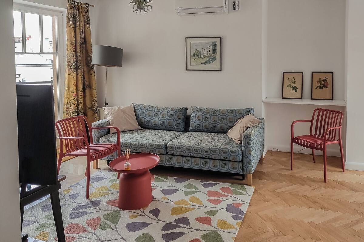 Un salon comprenant un canapé, deux fauteuils rouges, une petite table avec un pied rouge, un tapis coloré et un téléviseur.