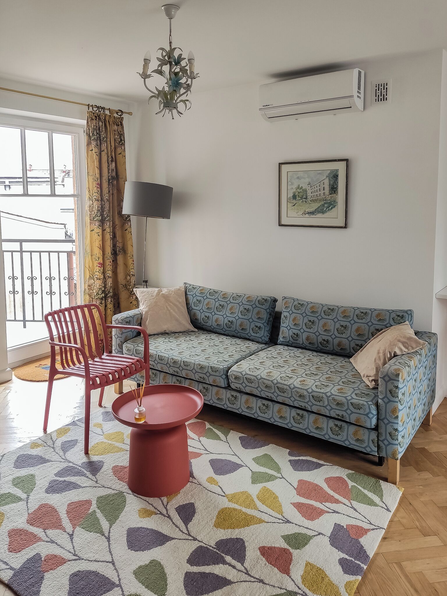 A living room with a patterned sofa, a red chair, a small round table, a chandelier, and a window with curtains.
