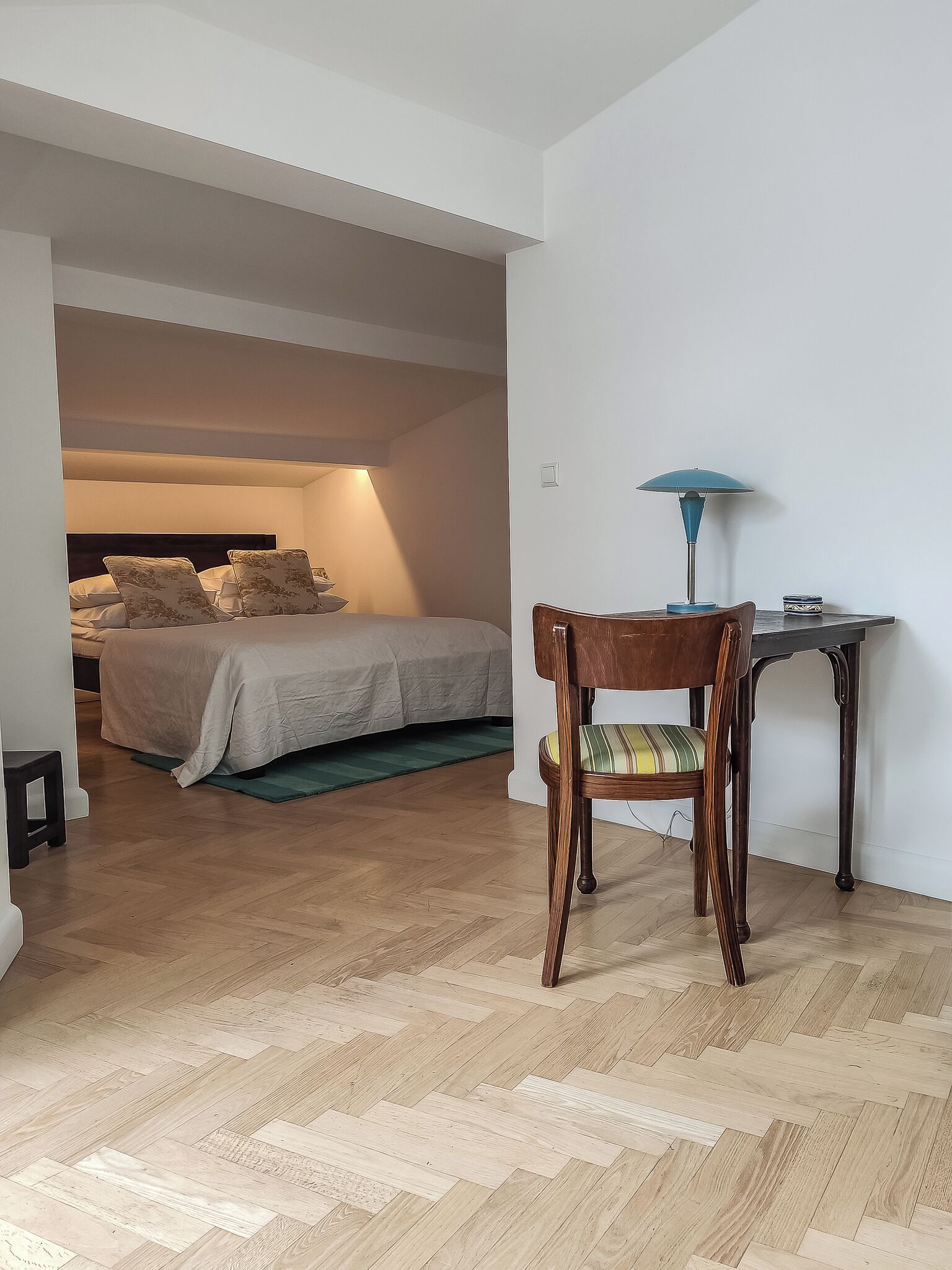 Quarto com cama, mesa de cabeceira e escrivaninha com abajur.
