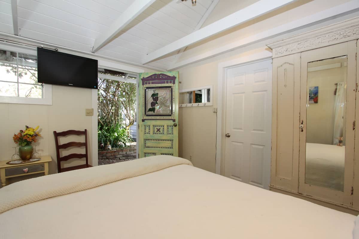 A bedroom with a bed, a TV mounted on the wall, a wooden chair, a small table with a vase of flowers, and a door with a mirror.