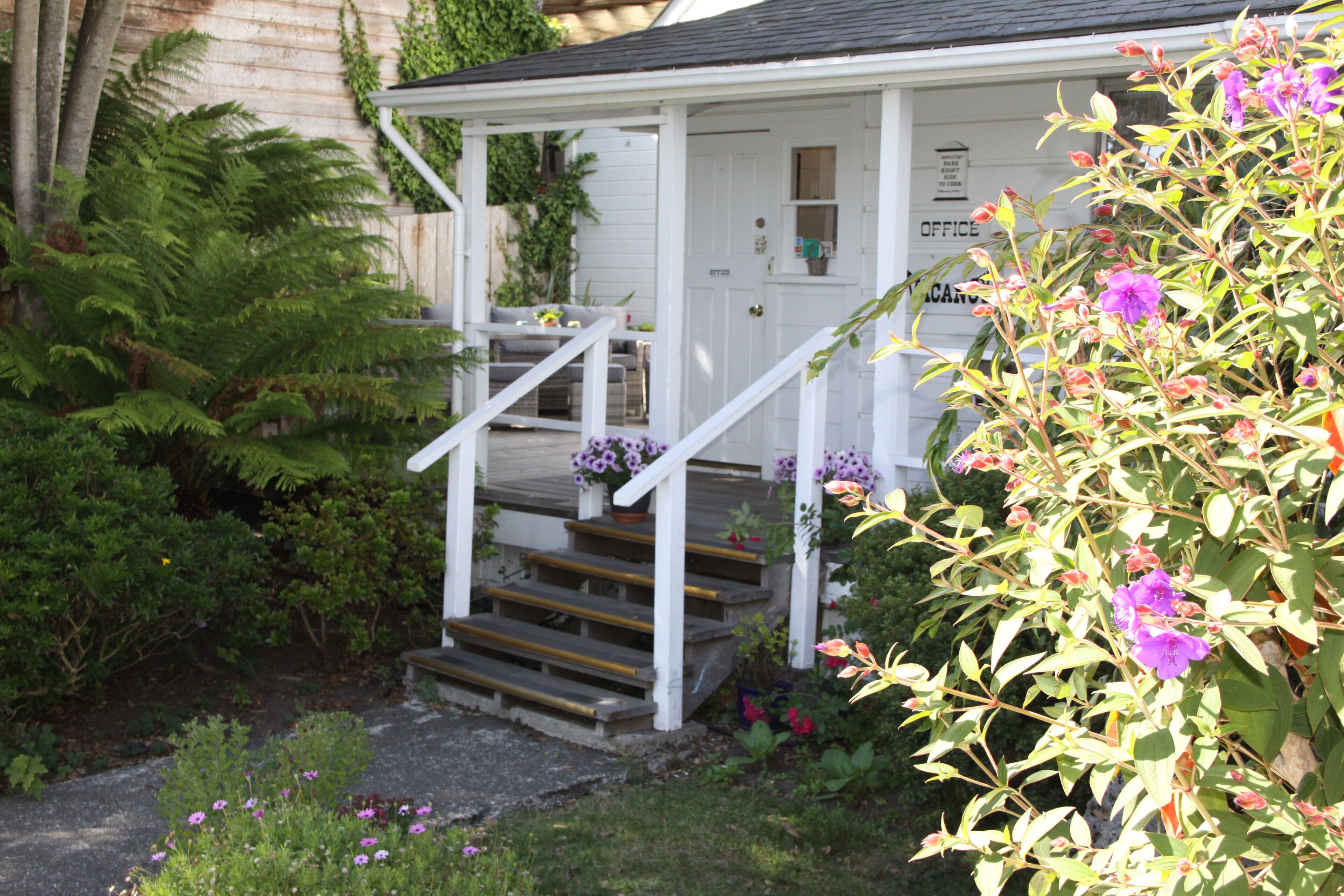 A white building with a porch, steps, and a garden with various plants and flowers.