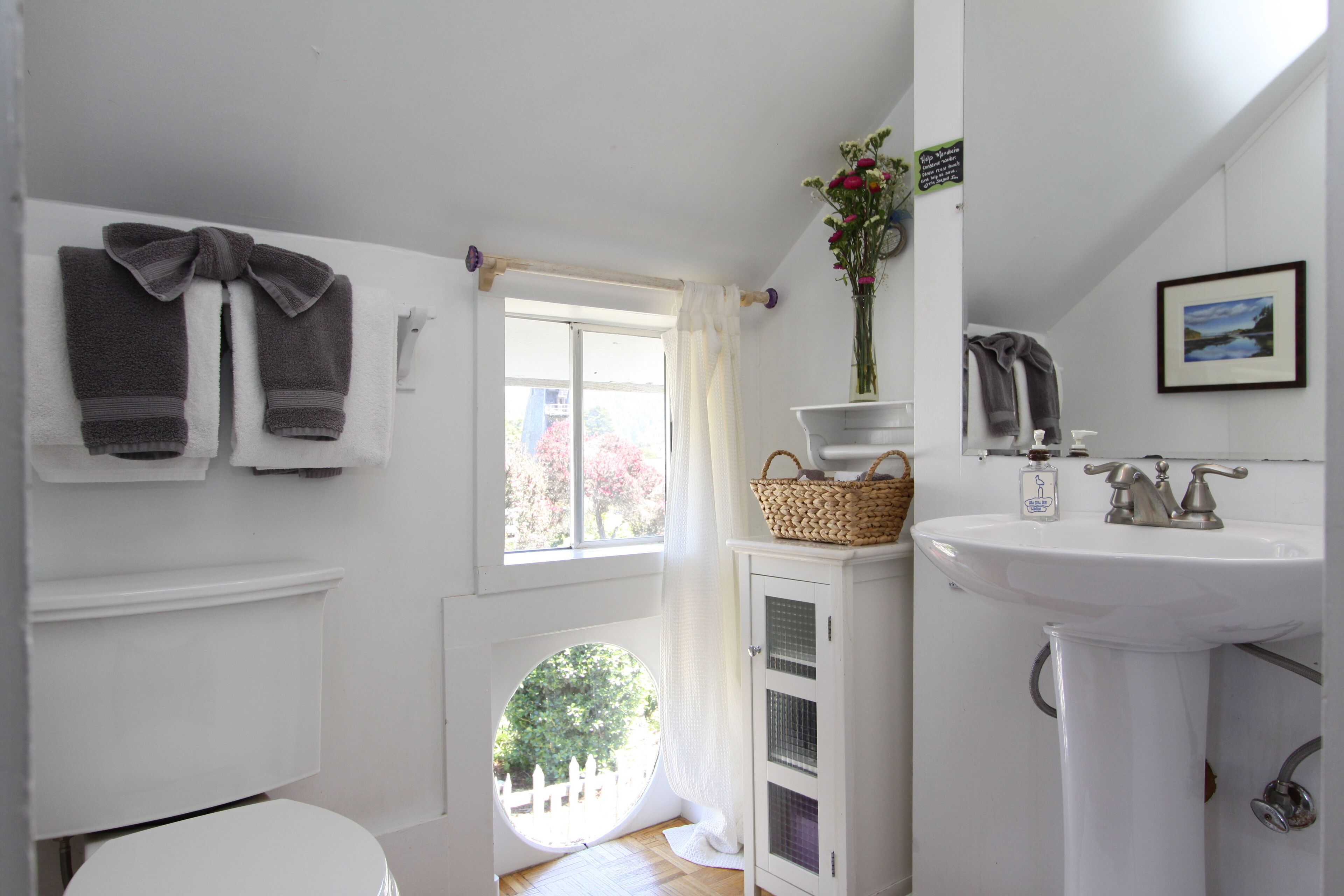 A bathroom with a white sink, a white toilet, a window with a view of trees, and towels hanging on the wall.