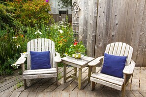 Two wooden Adirondack chairs with blue cushions, a small wooden table with a vase and glass, and a wooden fence in a garden setting.