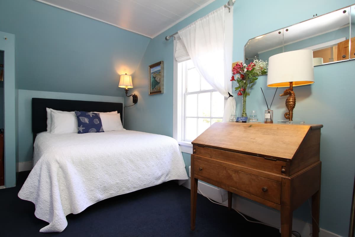 A bedroom with a bed, a wooden desk, a mirror, a window with white curtains, and a wall-mounted lamp.