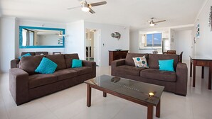 A spacious living room with two brown sofas, a glass coffee table, and ceiling fans.