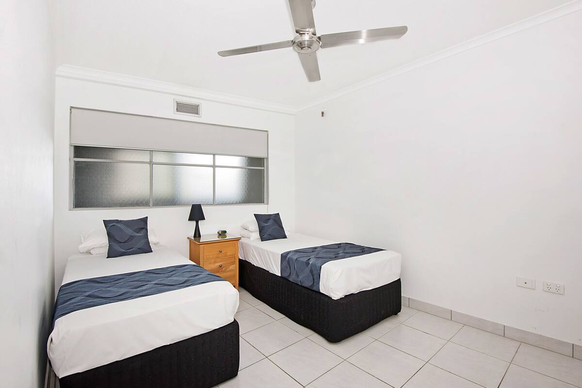 A hotel room with two single beds, a ceiling fan, a large window, and a wooden nightstand.
