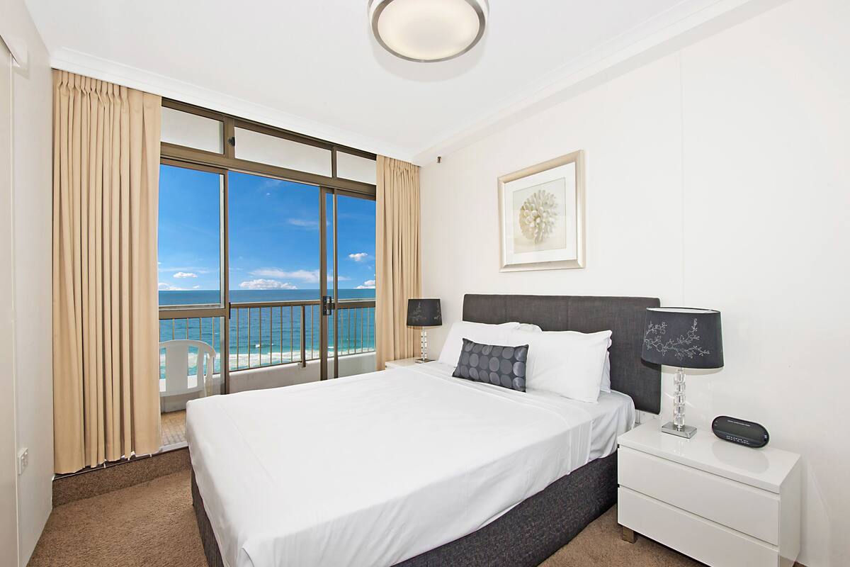 A hotel room with a large bed, a nightstand with a lamp, a framed picture on the wall, and a view of the ocean through a large window.