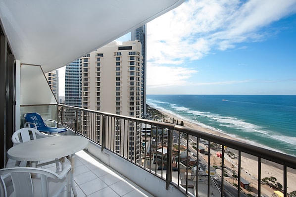 A balcony with a white table and chairs, overlooking a beach and ocean with high-rise buildings in the background.