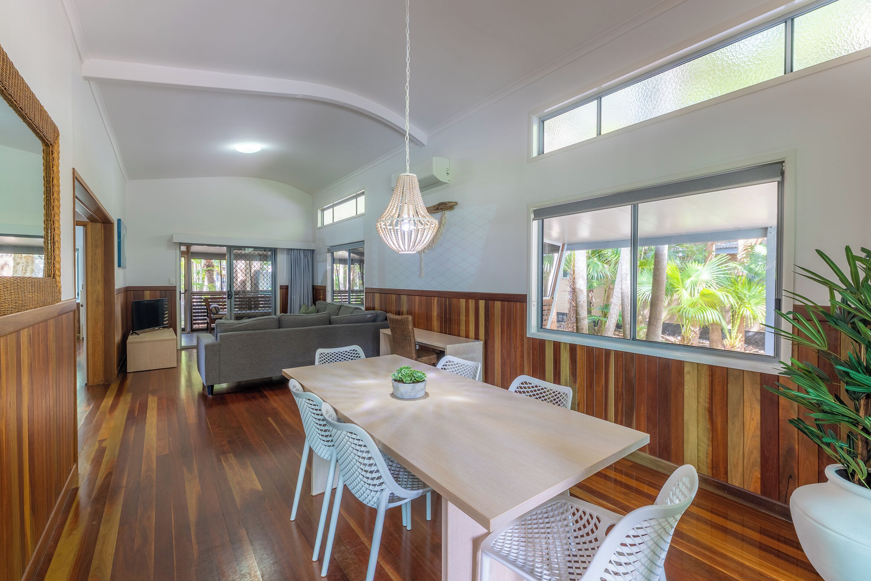 A modern dining area with a wooden table, white chairs, a hanging light fixture, and large windows with a view of greenery.