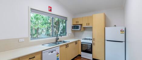 Cocina compacta con microondas, estufa y refrigerador, armarios de madera, lavavajillas y ventana con vista a zonas verdes.