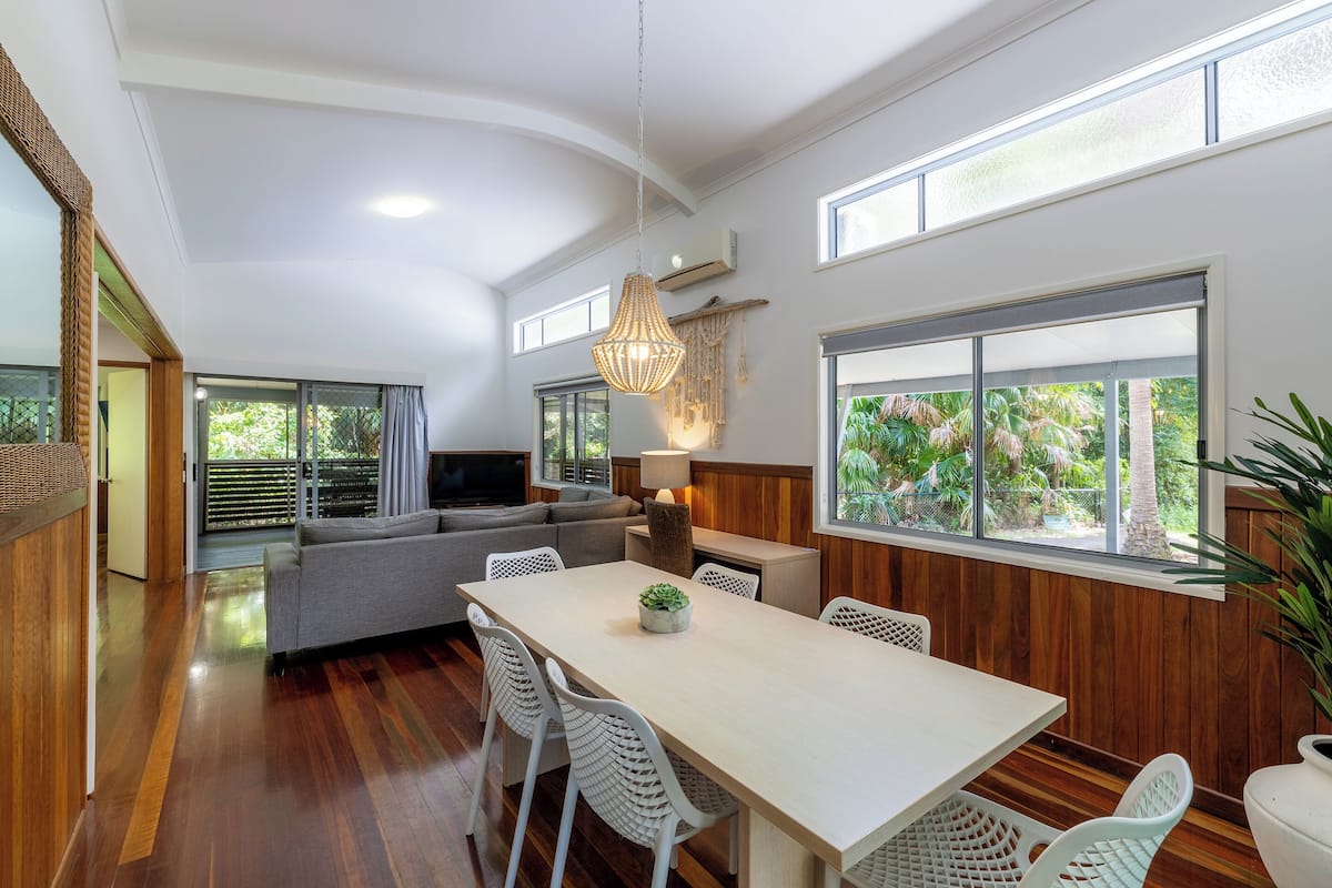 A spacious living area with a sofa, dining table, and chairs, a TV, and large windows.