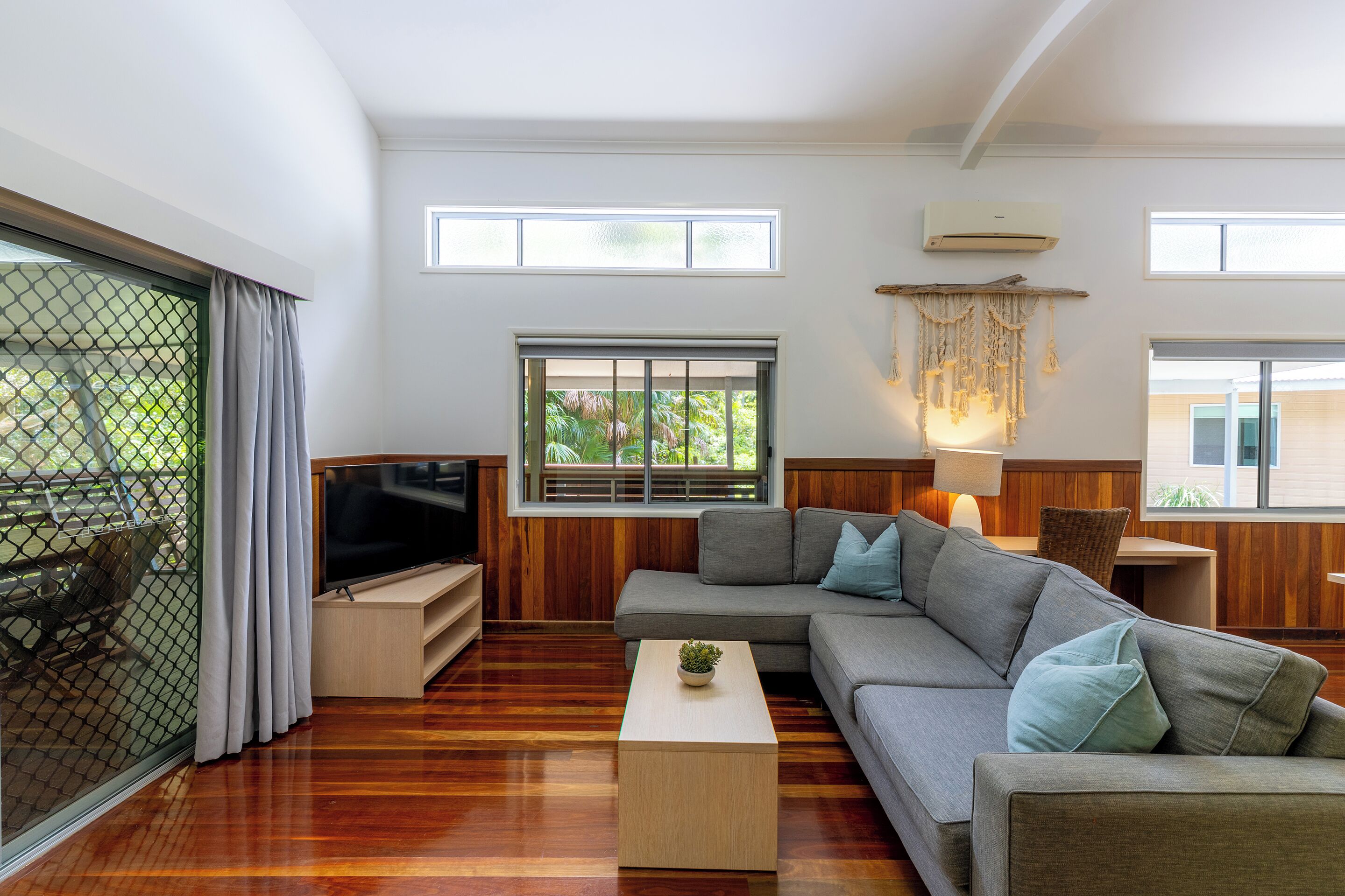 Un salon moderne avec un canapé d’angle, un téléviseur à écran plat, un plancher en bois et de grandes fenêtres.