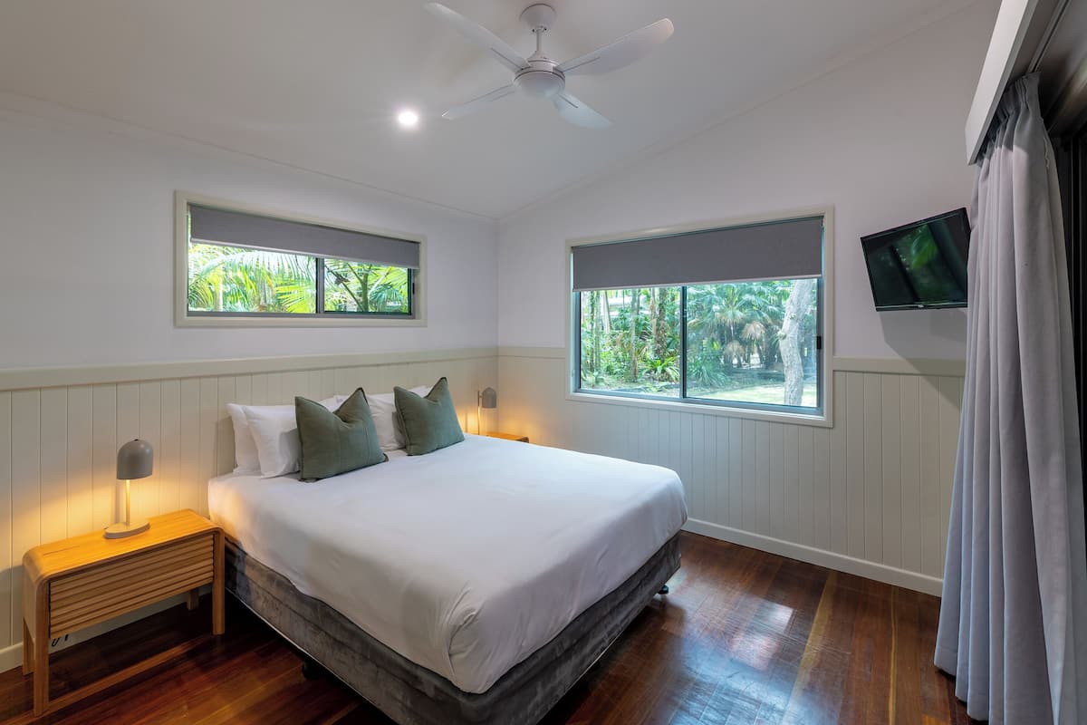 A bedroom with a bed, two bedside tables, a ceiling fan, two windows, and a television.