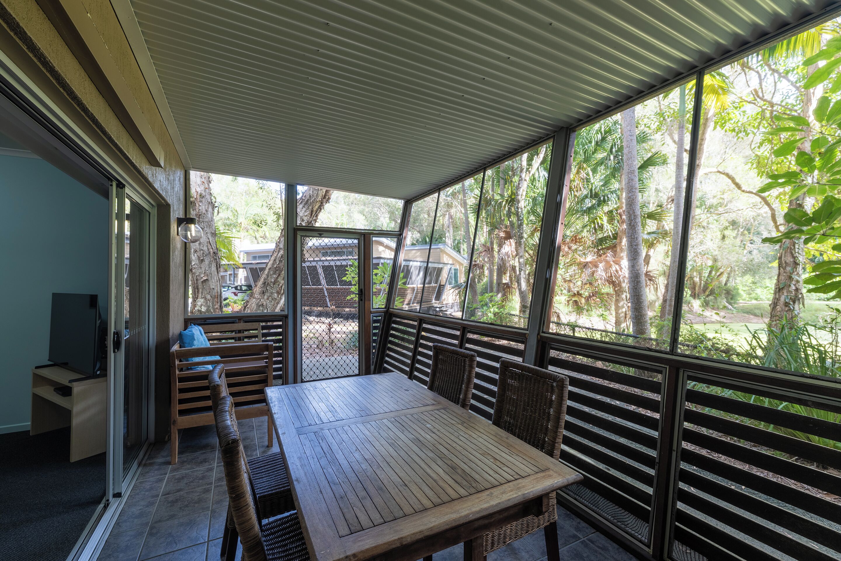 Un patio techado con una mesa y sillas de madera, un televisor sobre un mueble, y vistas al verdor exterior.